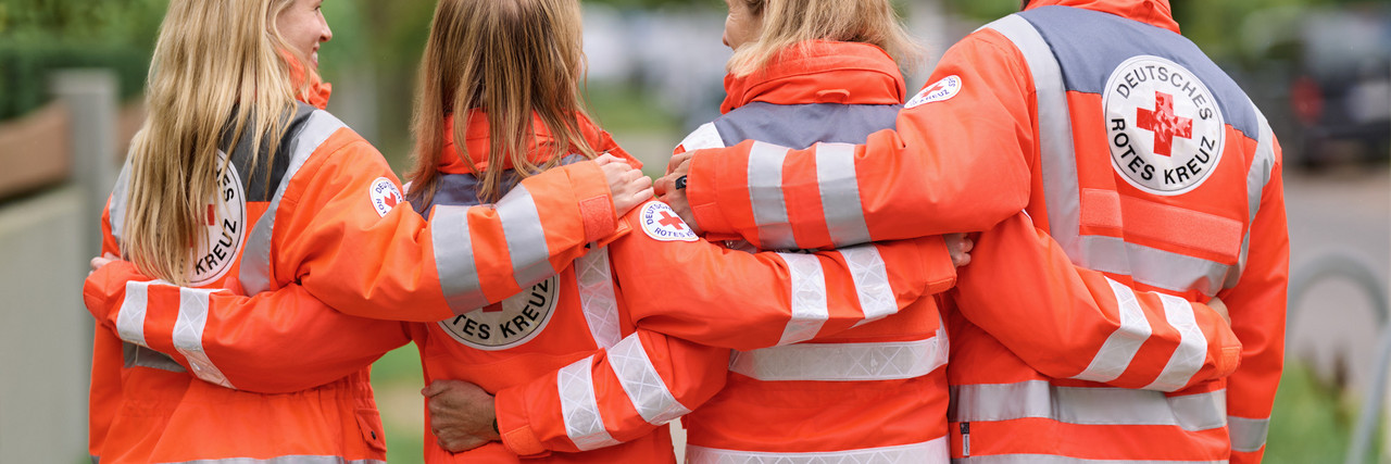 Vier Personen in orangefarbenen Jacken mit reflektierenden Streifen und dem Logo des Deutschen Roten Kreuzes stehen mit verschränkten Armen Rücken an Rücken im Freien