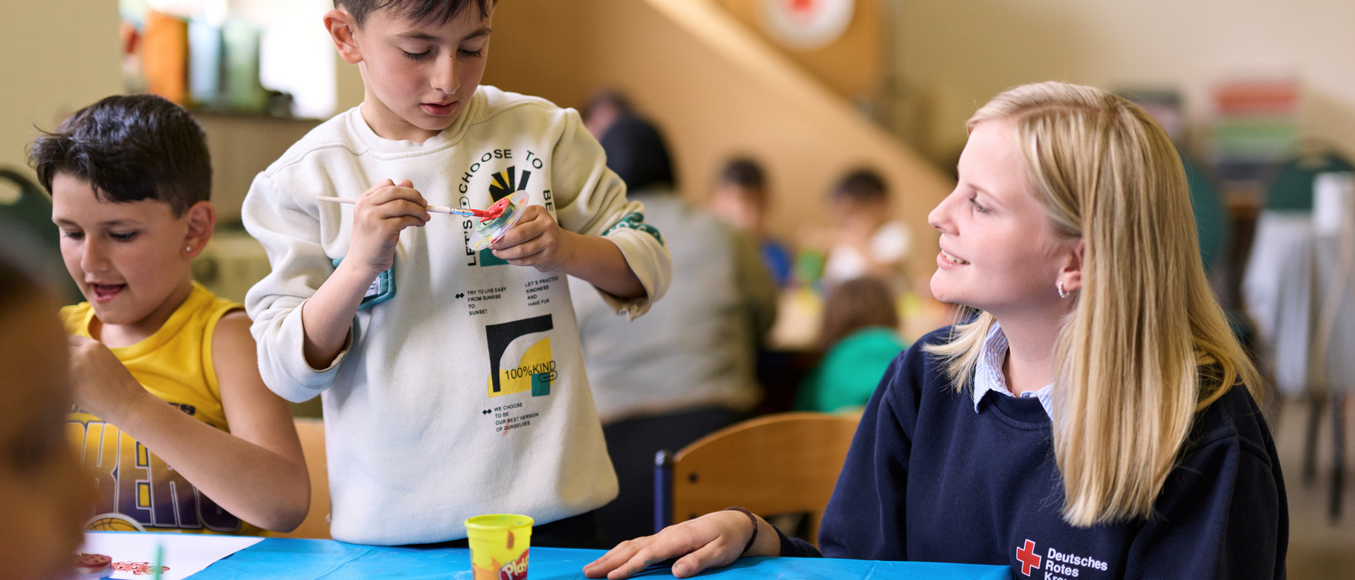 DRK-Freiwillige begleitet Kinder beim gemeinsamen Malen und Basteln an einem Tisch.