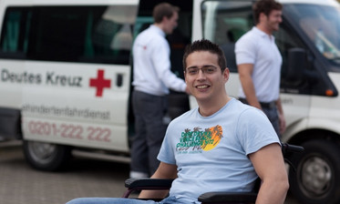 Junger Mann im Rollstuhl lächelt in die Kamera, im Hintergrund ein Fahrzeug und zwei Freiwillige vom DRK-Fahrdienst für Menschen mit Behinderungen